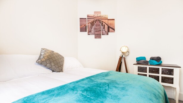 Un lit bien fait, recouvert d’une couverture turquoise, agrémenté d’un coussin à motifs et orné d’un tableau représentant une jetée, accroché au mur.