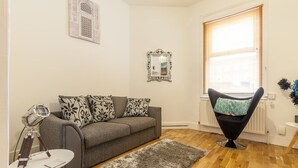 Un salon avec un canapé gris, des coussins à motifs, un fauteuil noir, une petite table blanche avec une lampe et un tapis gris.