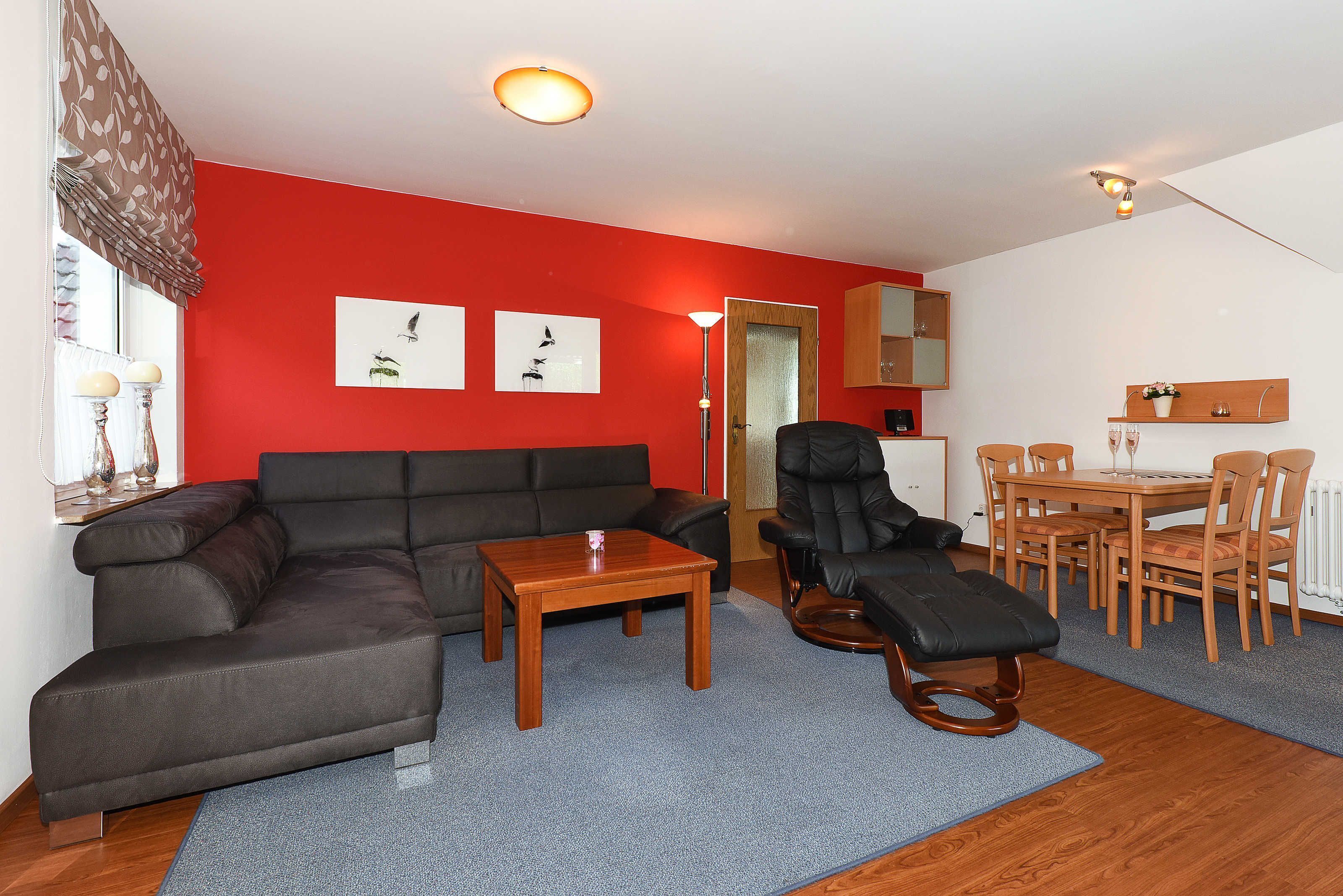 A living room with a red wall, a black leather sofa, a wooden coffee table, a black recliner, and a dining area with wooden furniture.