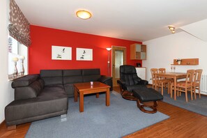 En stue med rød væg, en sort lædersofa, et træbord og en sort lænestol, samt et spiseområde med træmøbler.
