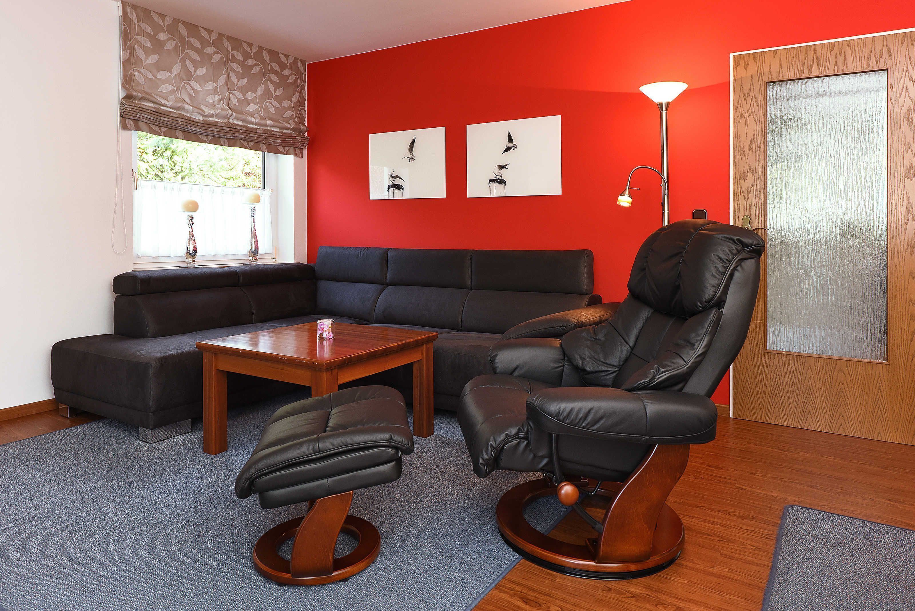 Un salon avec un mur rouge, un canapé en cuir noir, une table basse en bois, un fauteuil en cuir noir et deux cadres photos accrochés au mur.