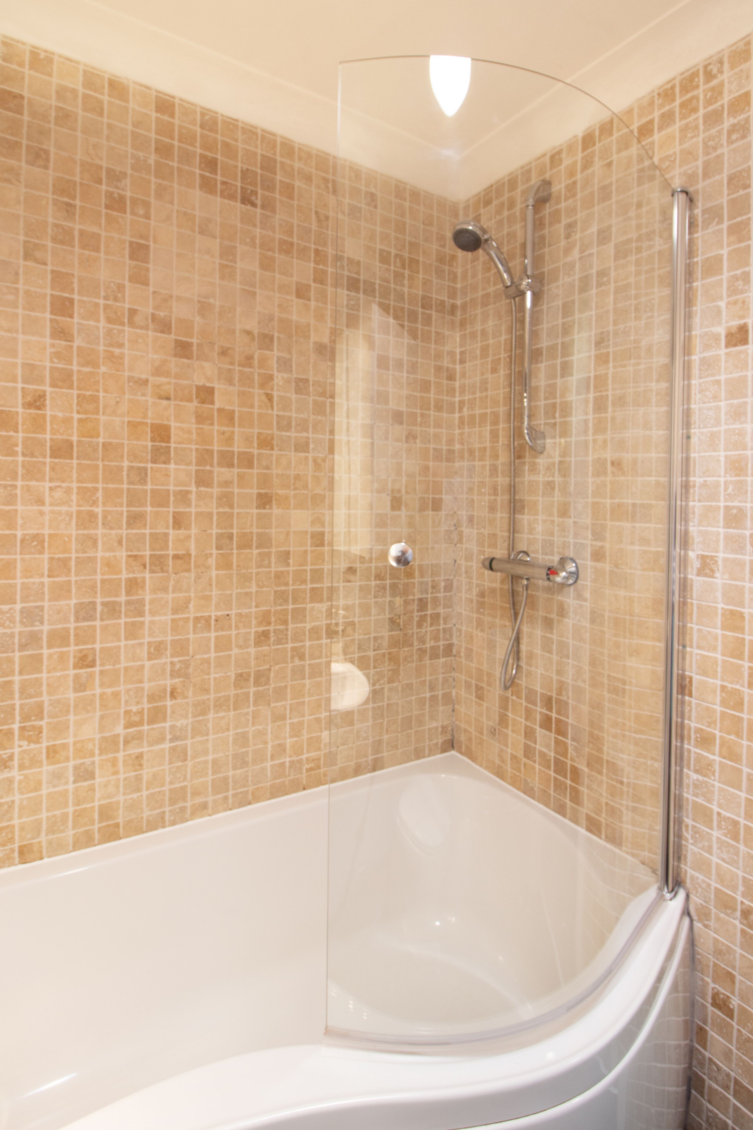 A modern bathroom with a glass-enclosed shower, a white bathtub, and beige mosaic tiles.