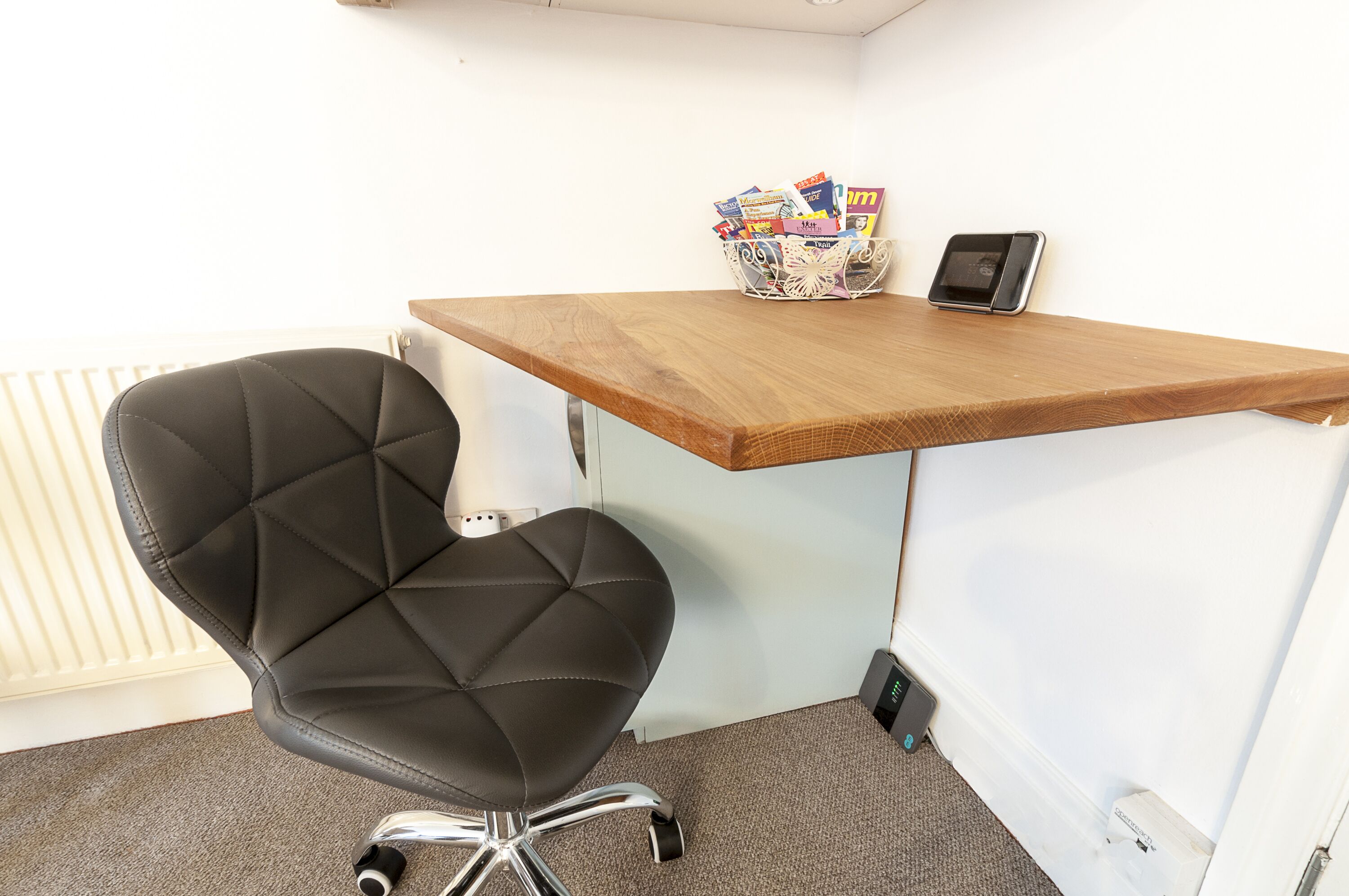 Een kleine kantoorruimte met een zwarte bureaustoel, een houten bureau en een mand vol tijdschriften.