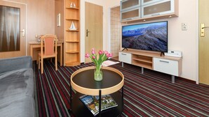 Moderni hotellihuone, jossa on televisio, pyöreä pöytä maljakko täynnä kukkia, sohva, kirjahylly ja raidallinen matto.