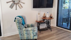 A living room with a patterned armchair, a round table with decorative items, a mounted TV, and a large mirror on the wall. - 2/2 condo daphne Al. (Daphne)