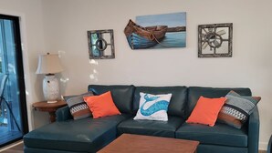 A living room with a dark green sofa, a wooden coffee table, and a ceiling fan. There are two framed pictures on the wall, a lamp on a side table, and a window with a view of the outdoors. - 2/2 condo daphne Al. (Daphne)