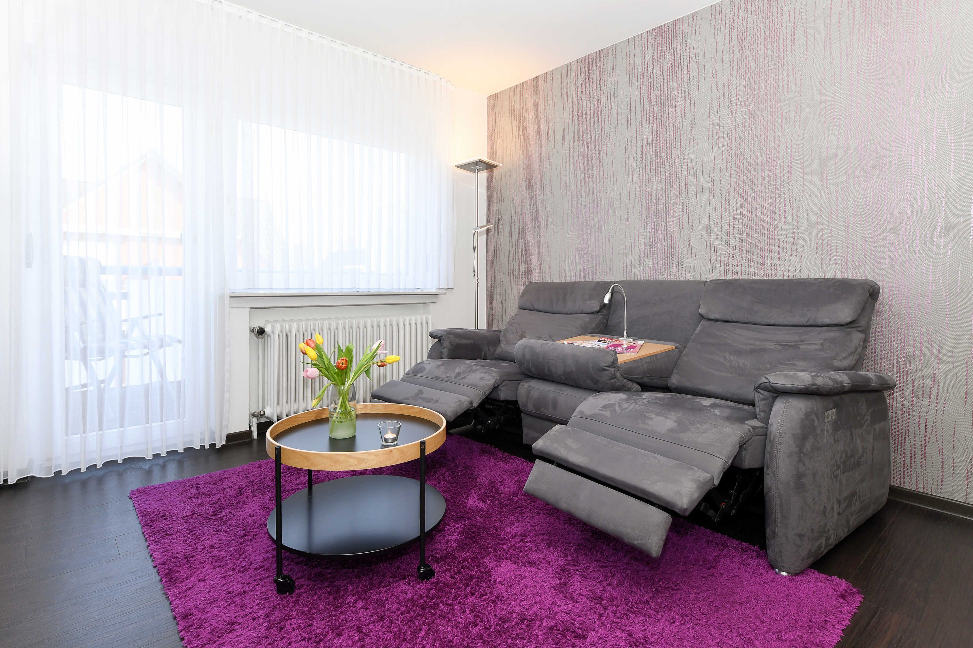 A modern living room with a grey reclining sofa, a purple rug, and a small round table with a vase of flowers.