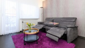 Un salon moderne avec un canapé gris inclinable, un tapis violet et une petite table ronde sur laquelle est posé un vase de fleurs.