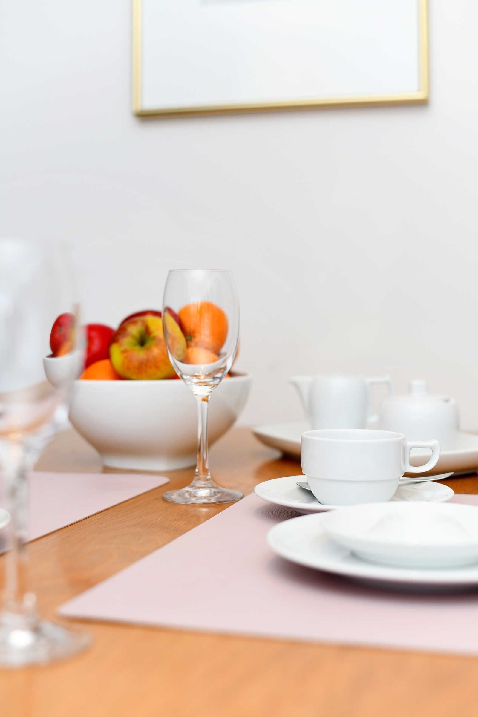 Un desayuno con un bol de fruta, una copa de vino y un juego de té blanco sobre una mesa de madera.