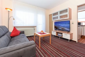 Quarto de hotel com sofá cinzento, almofada vermelha, mesa de madeira com uma tigela de fruta, televisão de ecrã plano, minibar e tapete riscado.