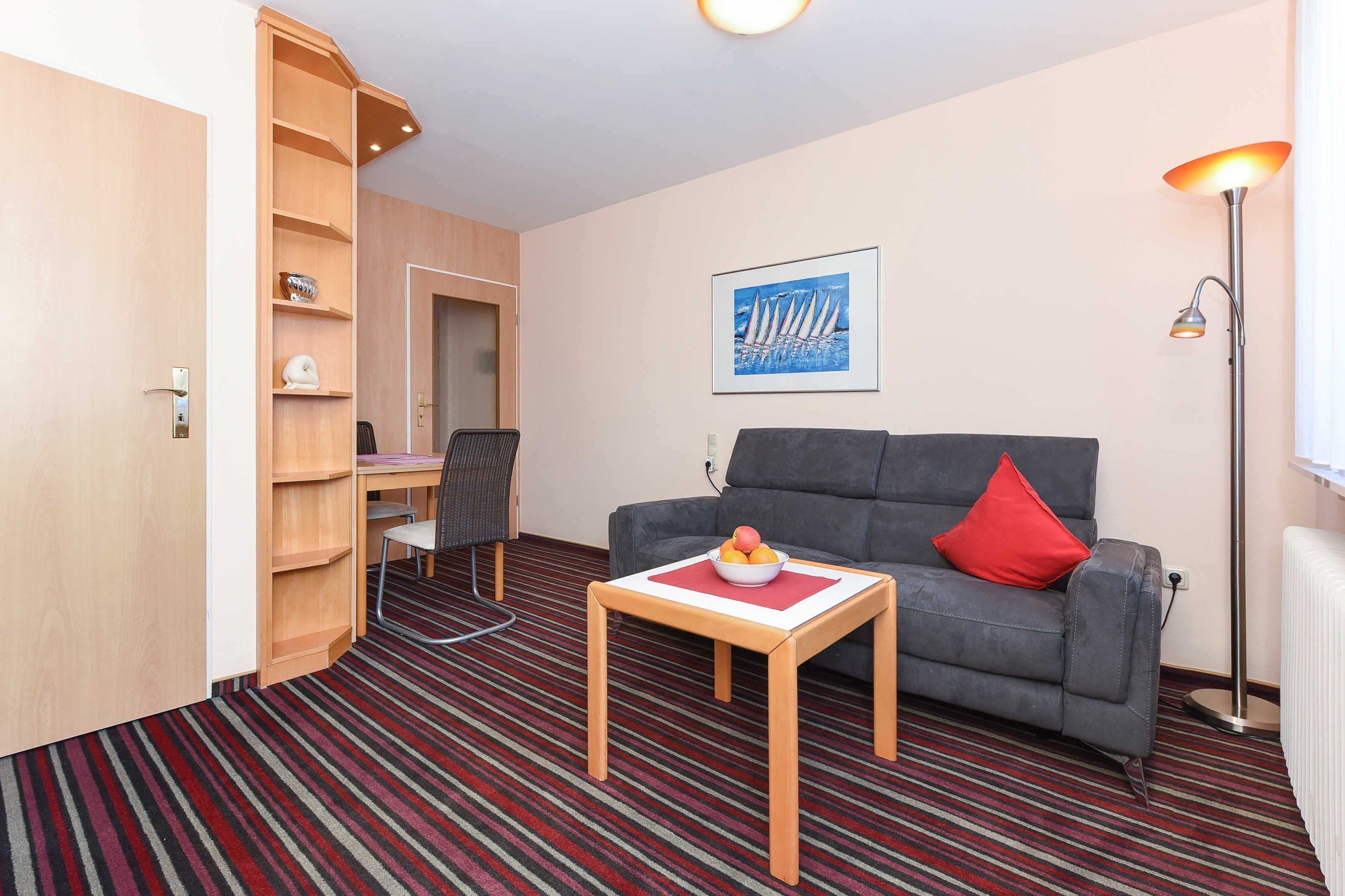 Una habitación moderna con un sofá gris, una pequeña mesa de madera con fruteros, una estantería, un escritorio con una silla y un cuadro en la pared.