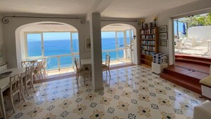 Una habitación amplia con un gran ventanal con vista al mar, un comedor con sillas y una estantería.