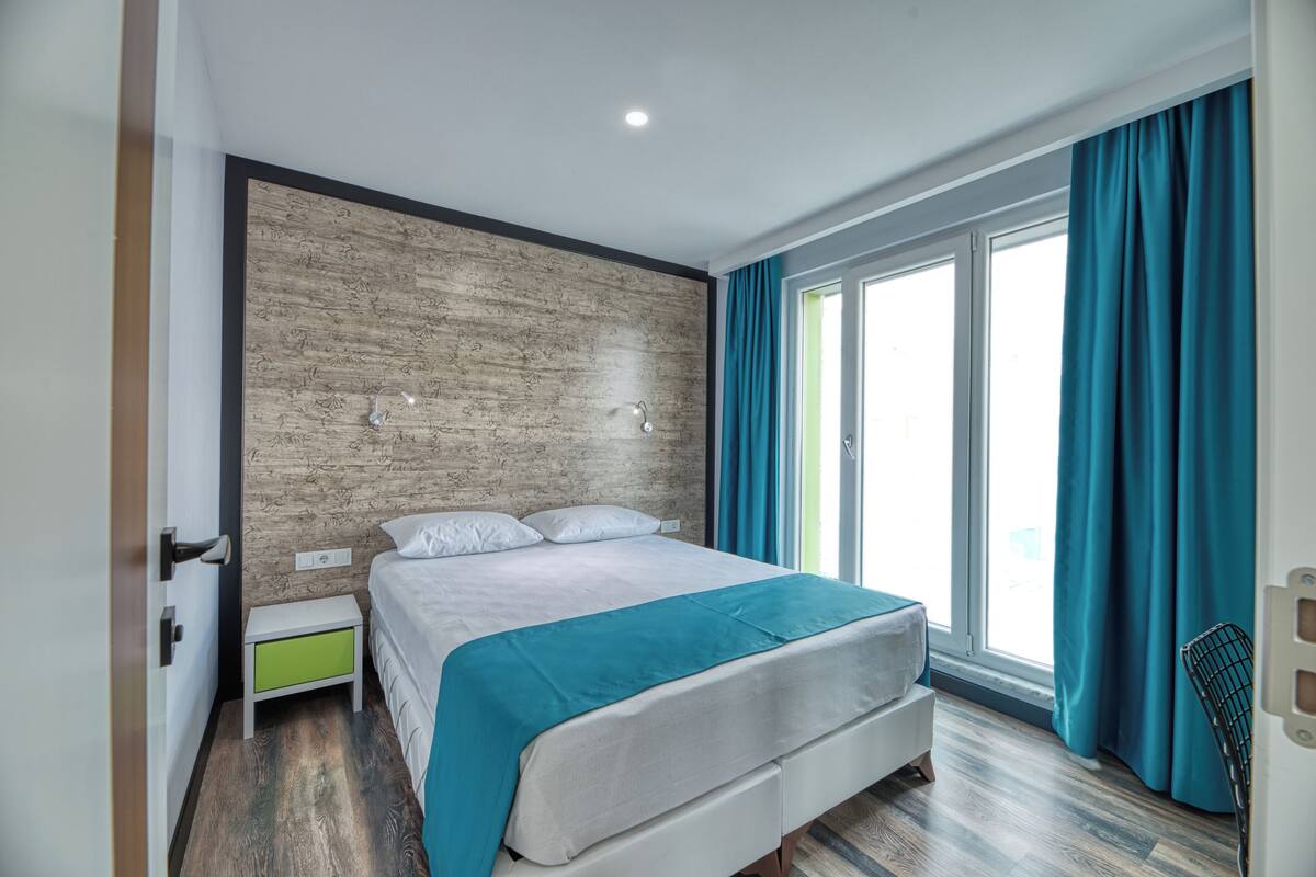 Une chambre d’hôtel moderne avec un grand lit, un mur décoratif en bois, une petite table de chevet et une vue sur l’extérieur grâce à une grande fenêtre.