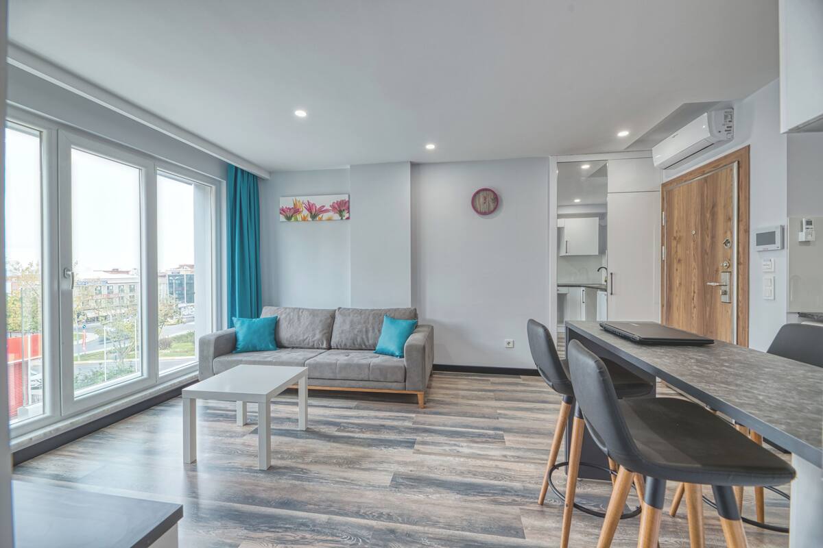 Un salon moderne avec un canapé gris, des coussins turquoise, une table basse blanche et un coin repas comprenant une longue table et des chaises noires.