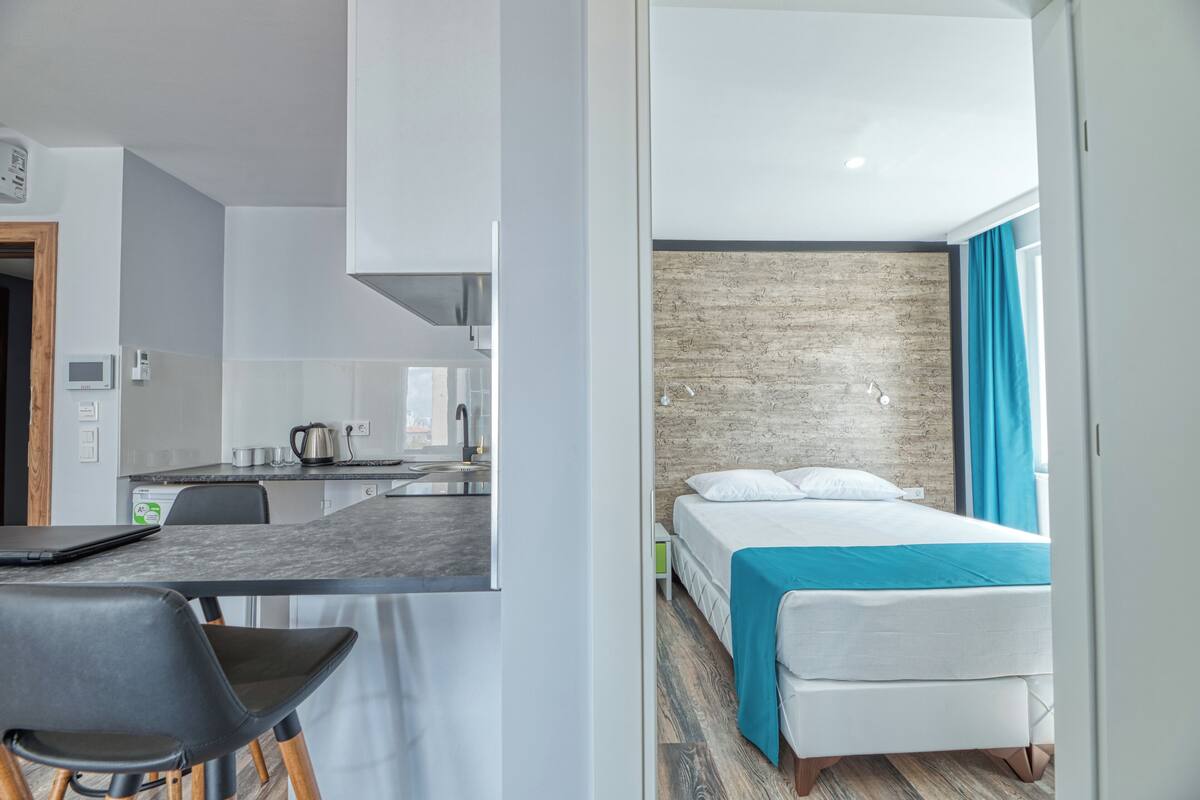 Une chambre d’hôtel moderne équipée d’un lit, d’un bureau avec une chaise, d’une kitchenette et offrant une vue sur la ville.