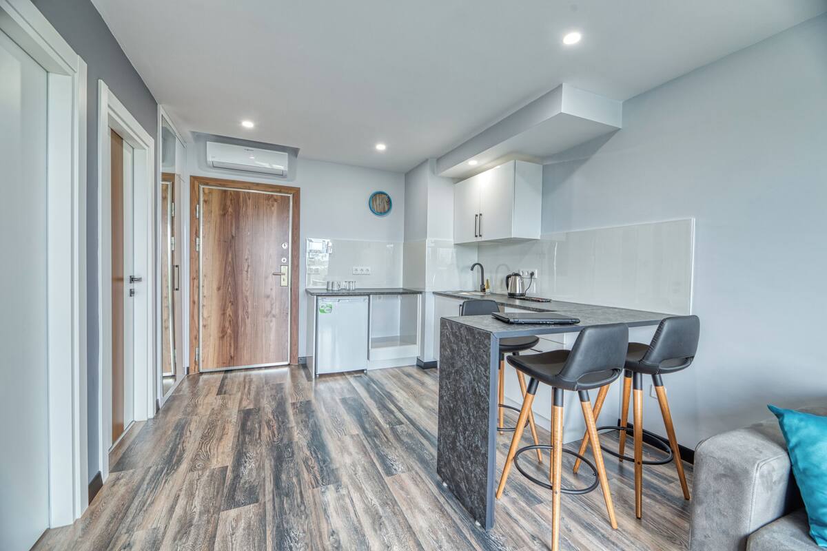Une cuisine moderne avec un îlot central, un plancher en bois et un canapé gris agrémenté d’un coussin turquoise.