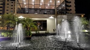 Un hôtel moderne avec une fontaine devant l’entrée, de grandes fenêtres à l’étage supérieur et un hall d’entrée bien éclairé.