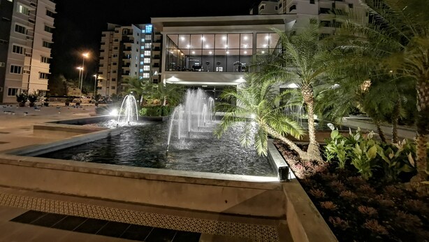 Une vue nocturne d’un bâtiment moderne avec une fontaine centrale, entouré de palmiers et d’une place bien éclairée.