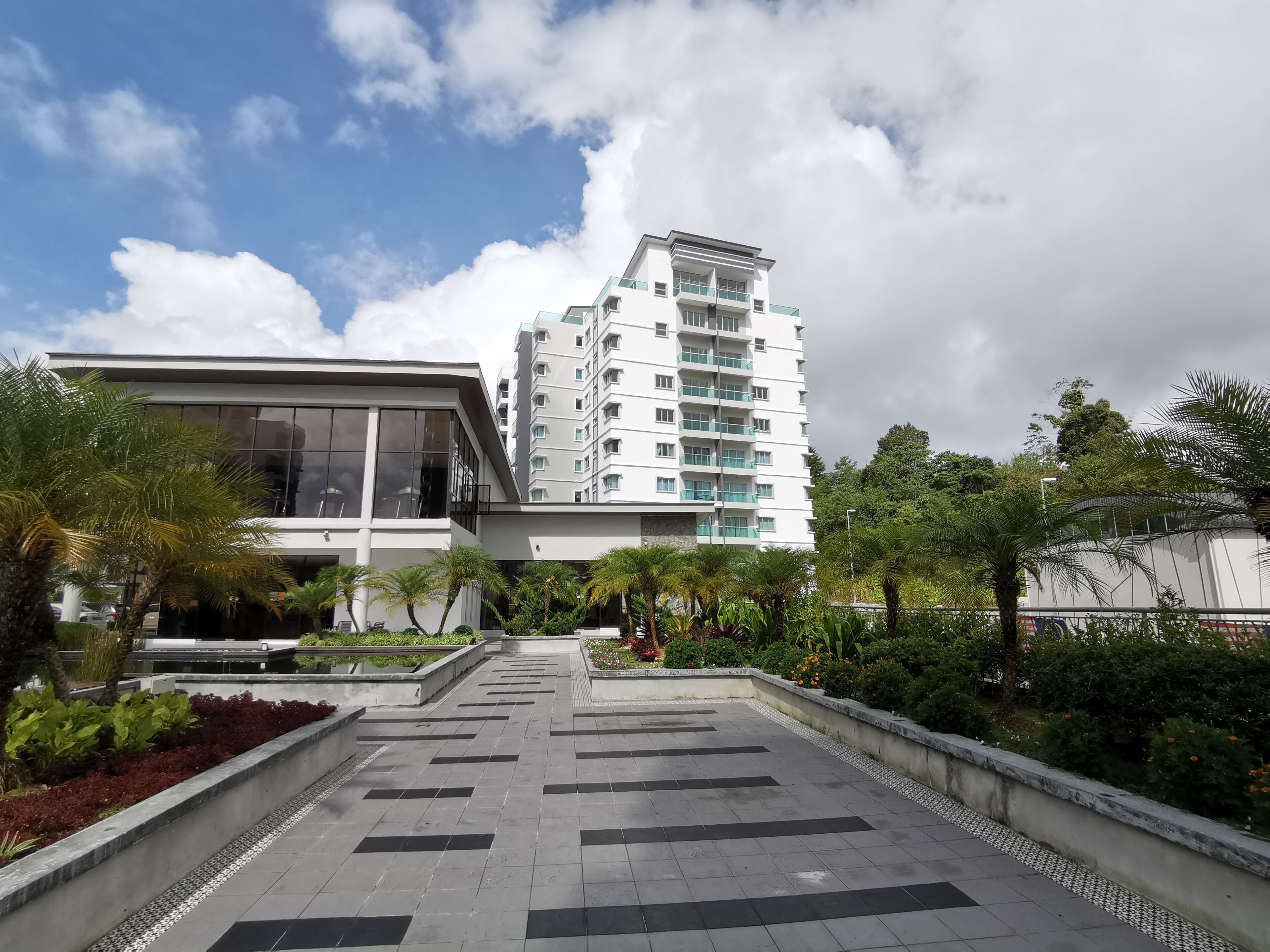 A modern building with a flat roof and large windows, surrounded by a landscaped area with palm trees and a paved walkway.