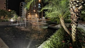 Une scène nocturne mettant en scène un élément aquatique avec des fontaines, entouré de palmiers et d’un bâtiment dont les fenêtres sont illuminées en arrière-plan.