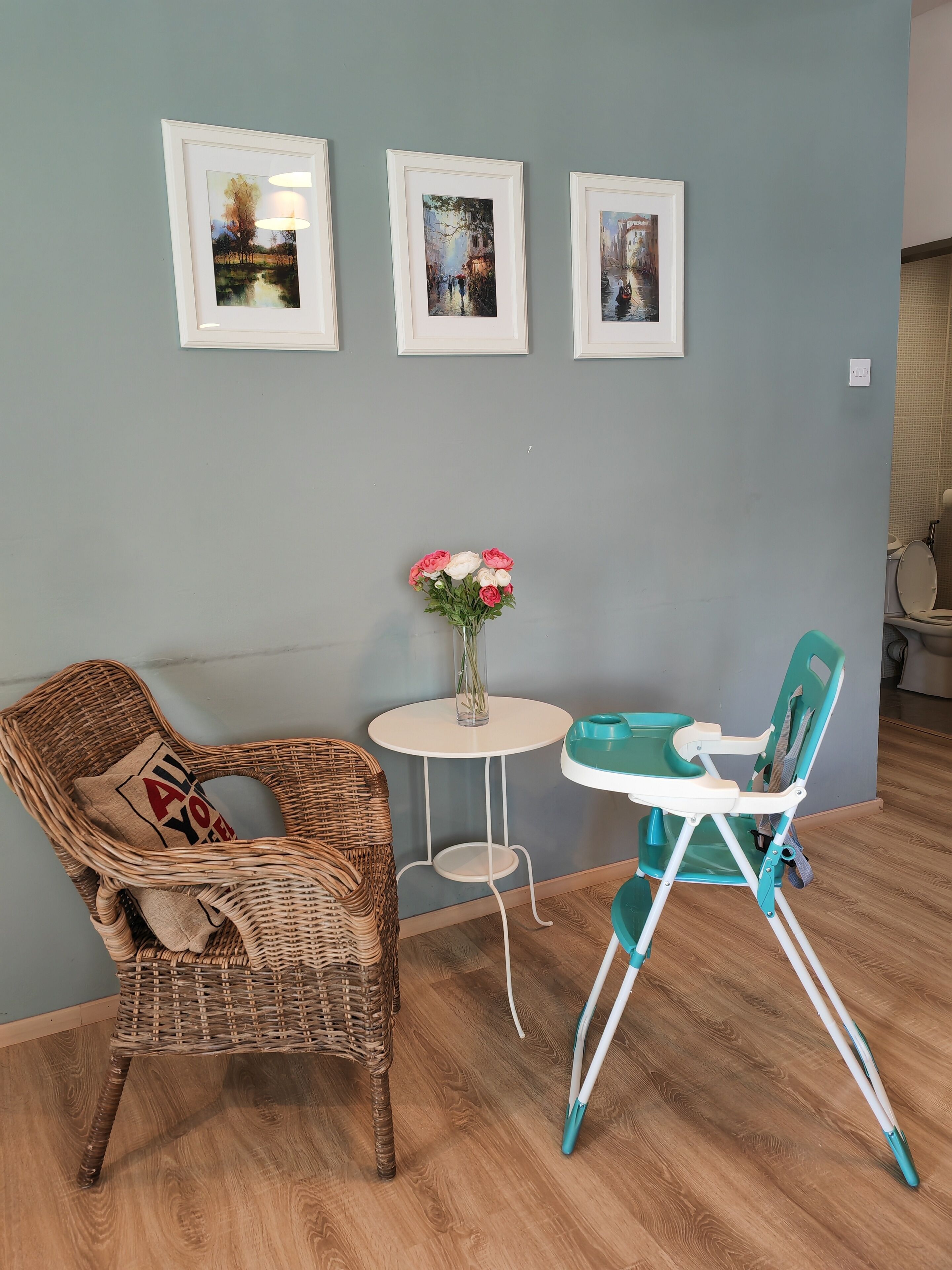 A room with a wicker chair, a small table with a vase of flowers, and a high chair. There are three framed pictures on the wall.