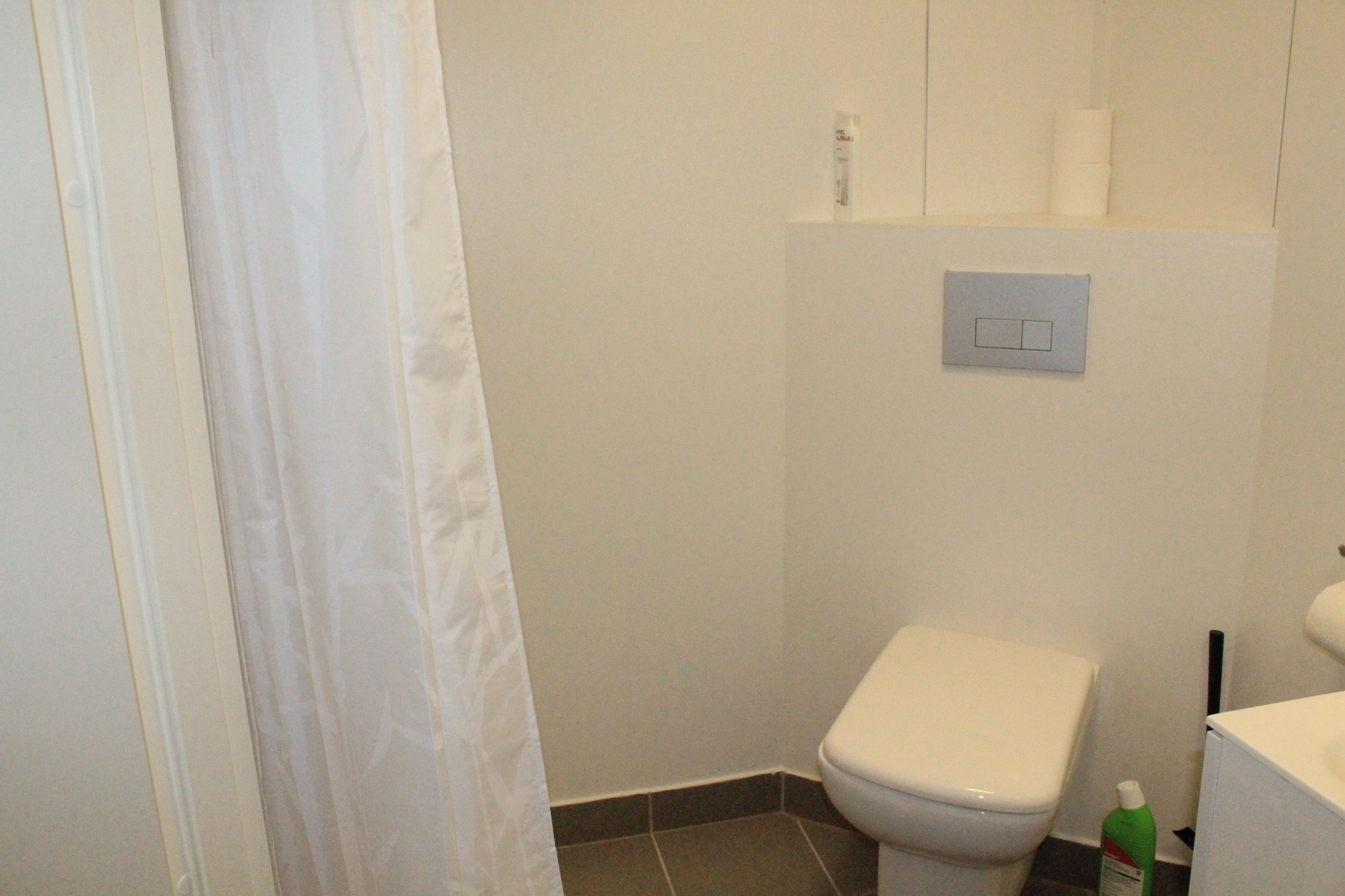 A bathroom with a toilet, a shower curtain, and a green bottle of cleaning product on the floor.
