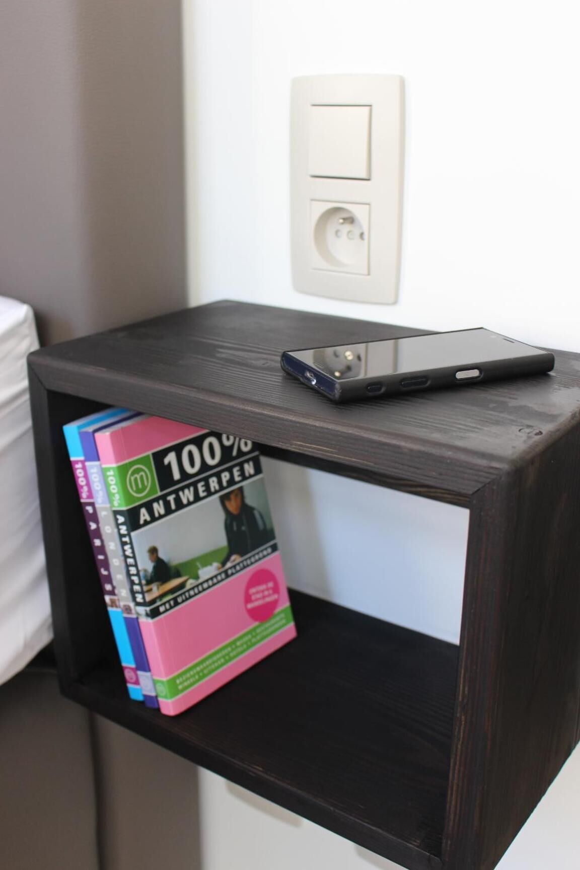 Un téléphone intelligent posé sur une table de chevet en bois sombre, avec un livre intitulé « 100 % Anvers » placé en dessous.