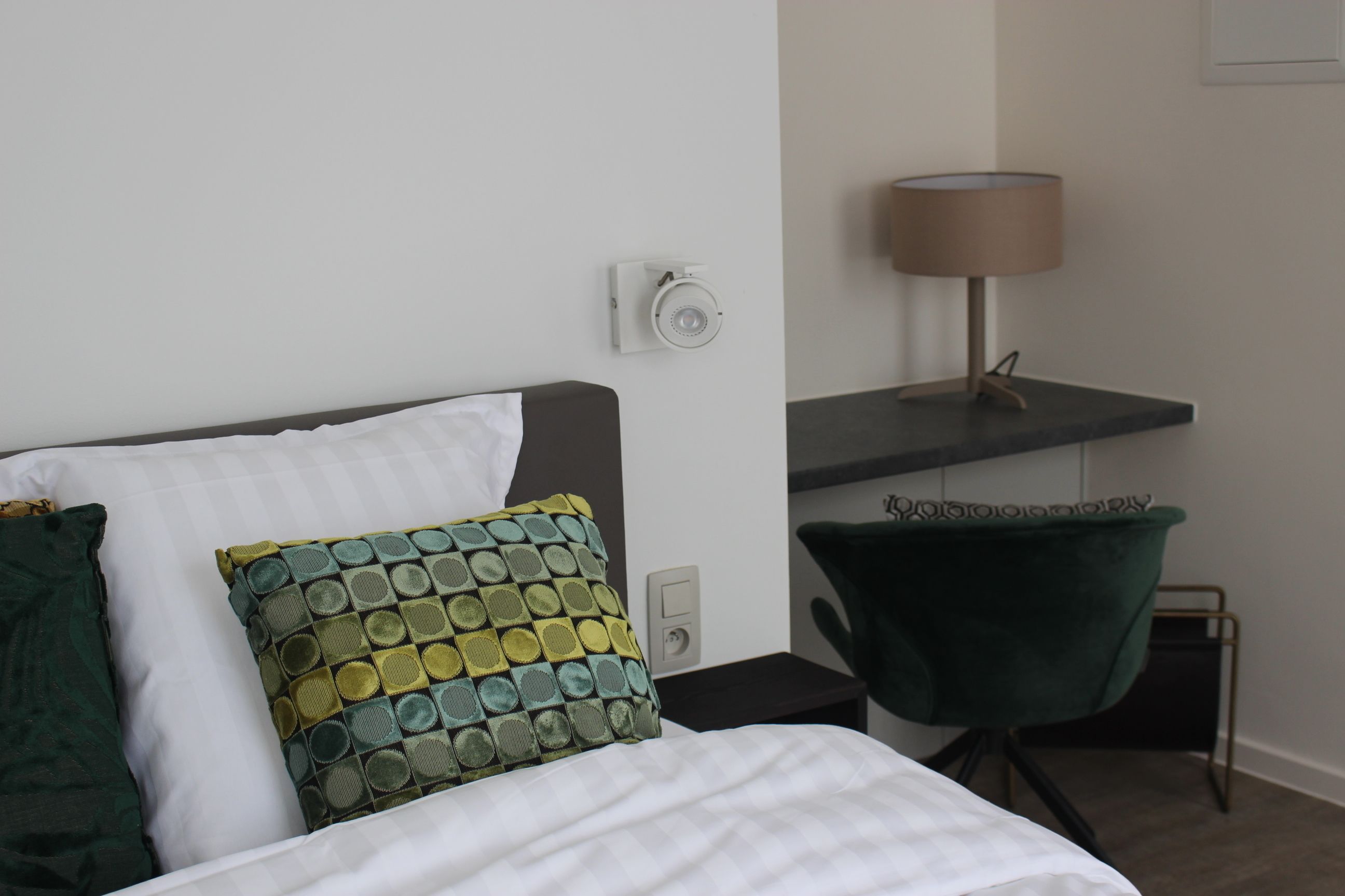 Un letto rifatto con cuscini bianchi e verdi, una scrivania con una lampada e una sedia verde in una stanza moderna.