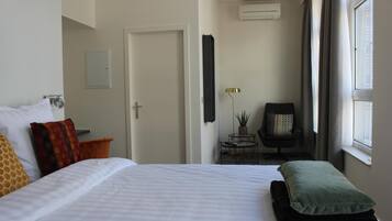 Habitación de hotel con cama, almohadas, una mesita con lámpara, una silla y una ventana con cortinas.