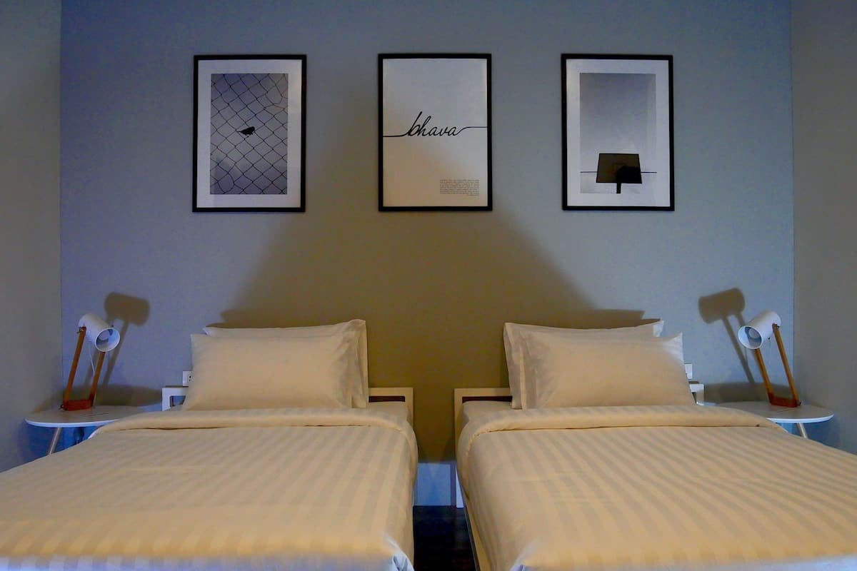 Ein Hotelzimmer mit zwei Betten, einem gerahmten Kunstwerk über jedem Bett und einem Nachttisch mit einer Lampe auf jeder Seite.