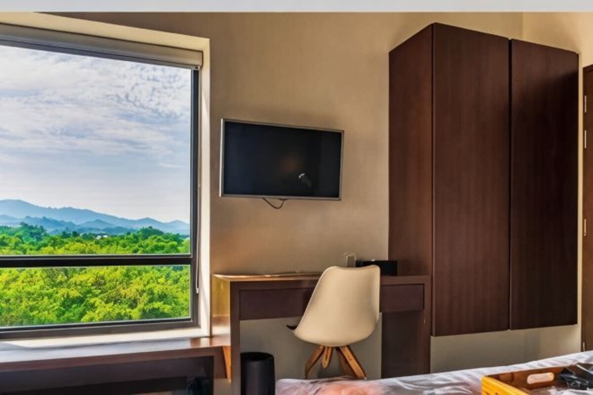 A room with a desk, chair, TV, wardrobe, and a window with a view of greenery and mountains.
