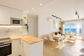 A modern kitchen with a built-in microwave, a dining area with a wooden table and chairs, and a balcony with a view of the sea.