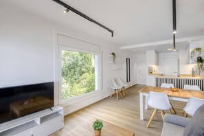 A modern living room with a flat-screen TV, a dining area with a wooden table and chairs, and a kitchen area with a countertop and sink.
