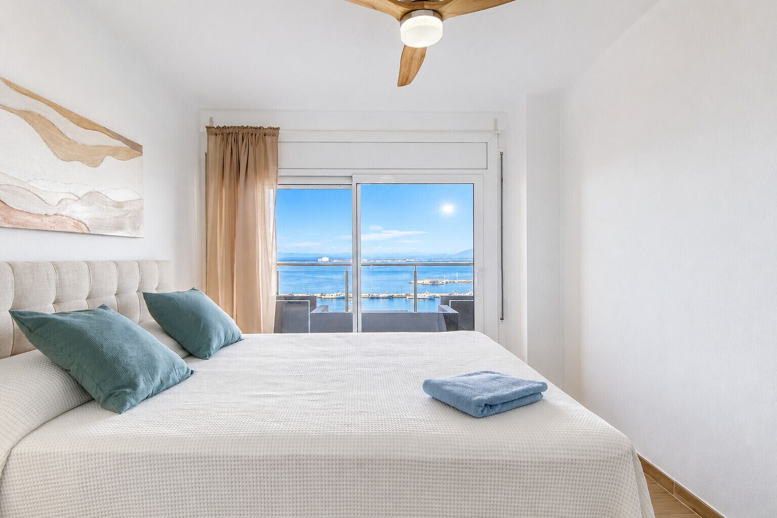 A bedroom with a large bed, a ceiling fan, a view of the sea, and a balcony.