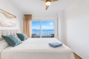 A bedroom with a large bed, a ceiling fan, a view of the sea, and a balcony.