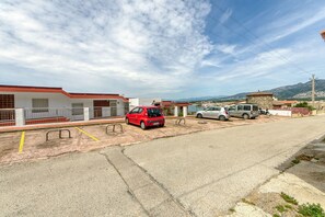 A parking area with a red car, a silver car, and a motorcycle. There are buildings with red roofs and white walls in the background.