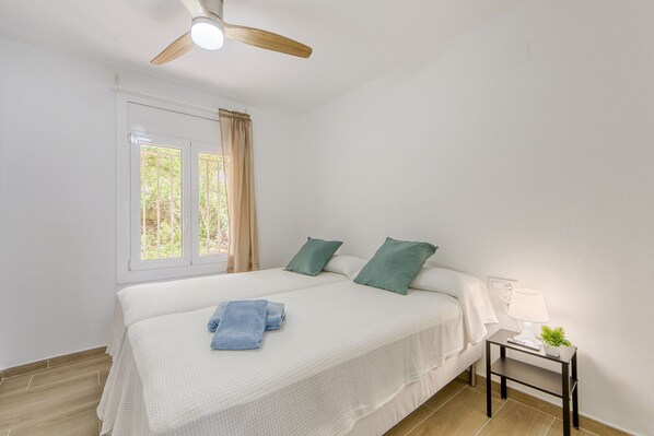 A bedroom with a bed, a window, a ceiling fan, a bedside table with a lamp, and a small plant.