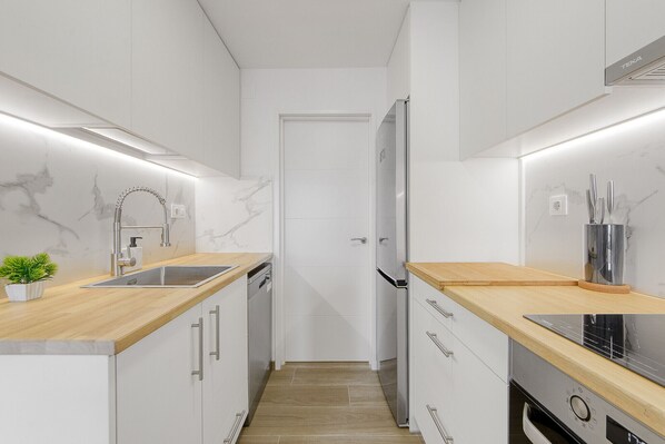 A narrow kitchen with white cabinets, a stainless steel refrigerator, and a wooden countertop.