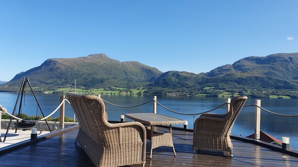 Une terrasse dotée de meubles en osier et d’une table, offrant une vue sur un lac avec des montagnes en arrière-plan.