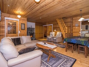 A cozy cabin interior with a wooden ceiling, a fireplace, a sofa, a dining table, and a staircase.