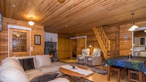 Een gezellig interieur van een chalet met een open haard, een bank, een eettafel en een trap.