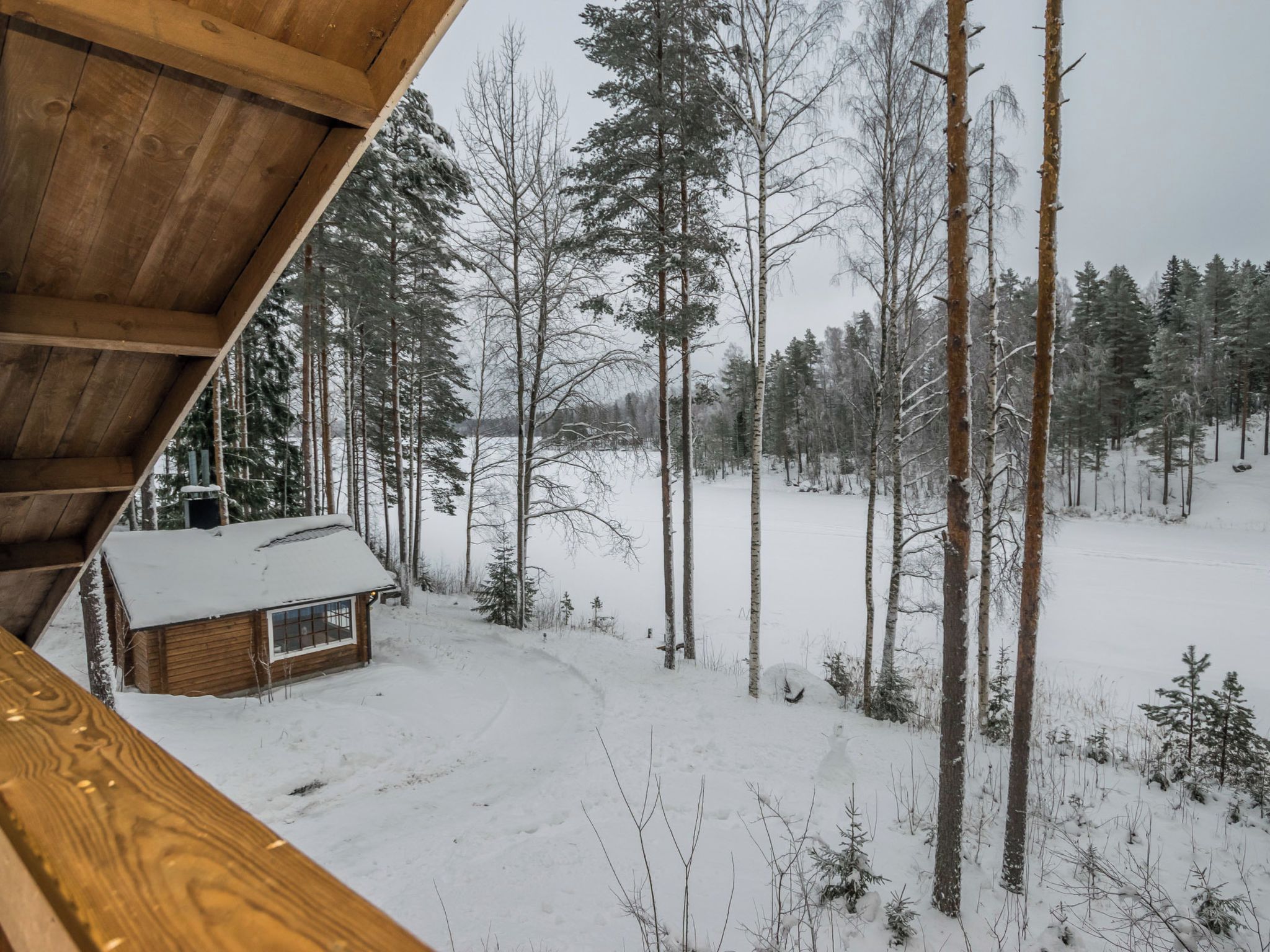 Una cabaña con techo cubierto de nieve, rodeada por un bosque de árboles altos y sin hojas.