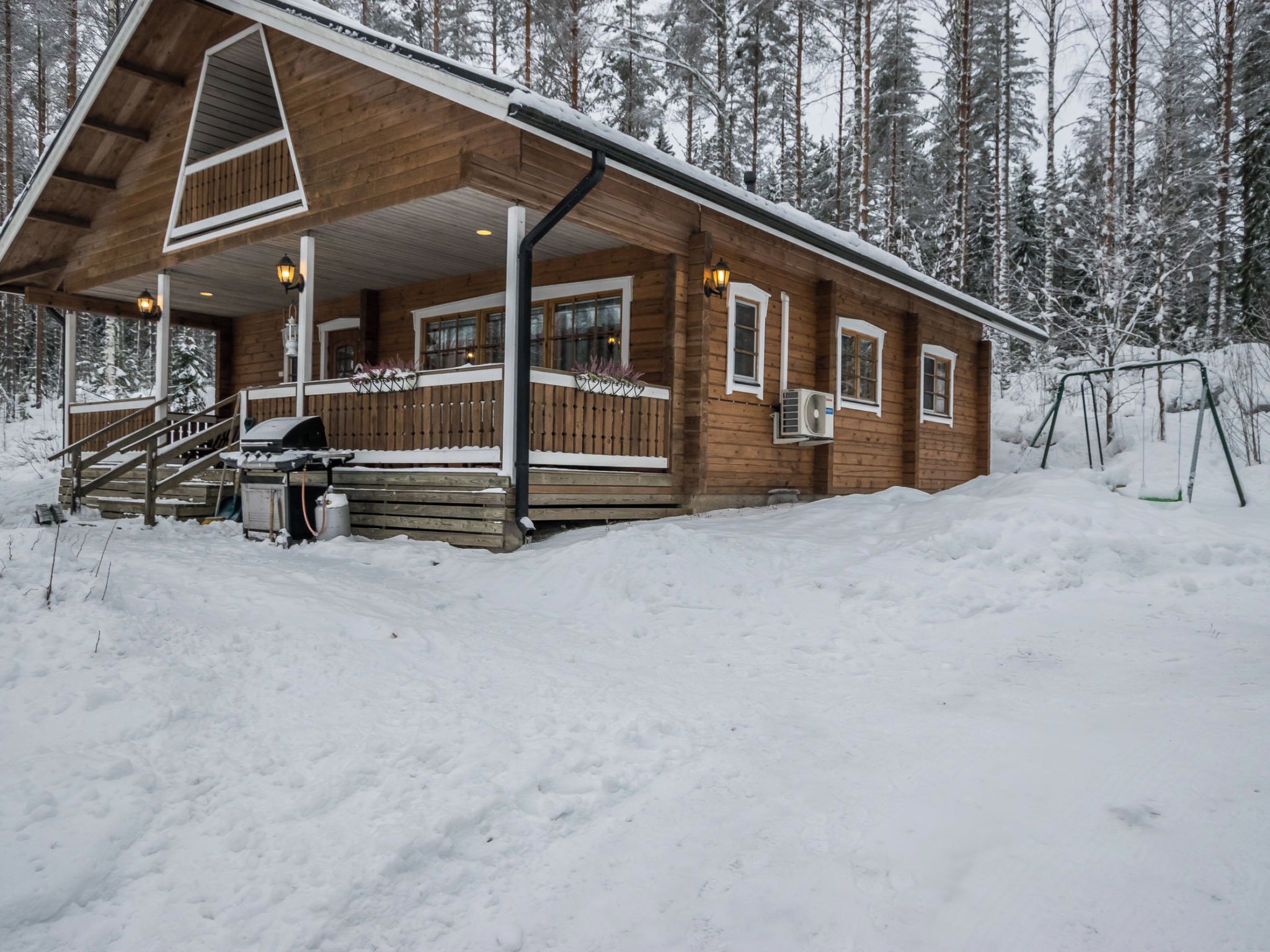 A wooden cabin with a covered porch, a grill, and a swing set in a snowy forest.