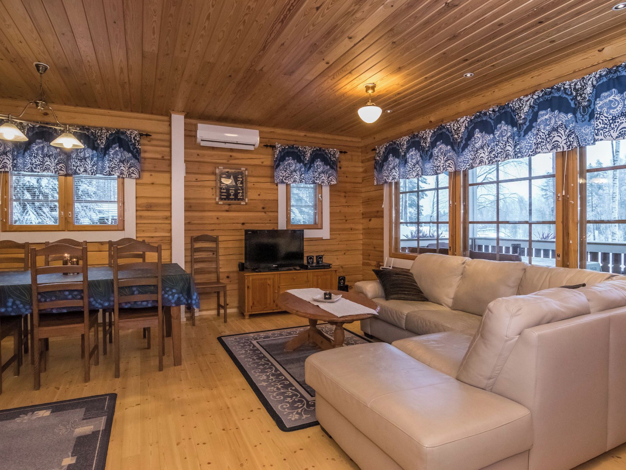 A cozy cabin interior with a wooden ceiling, a sofa, a TV, and a dining area.