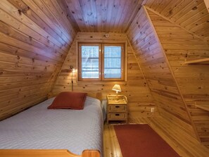 A small, cozy room with a sloped ceiling, a single bed, a nightstand with a lamp, and a window with blinds.