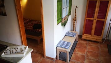 Quarto pequeno com uma cama, um banco, um armário e uma porta para outro cômodo.