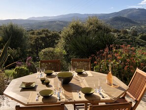 A wooden table set outdoors with bowls, glasses, and cutlery, surrounded by chairs, with a mountainous landscape in the background.