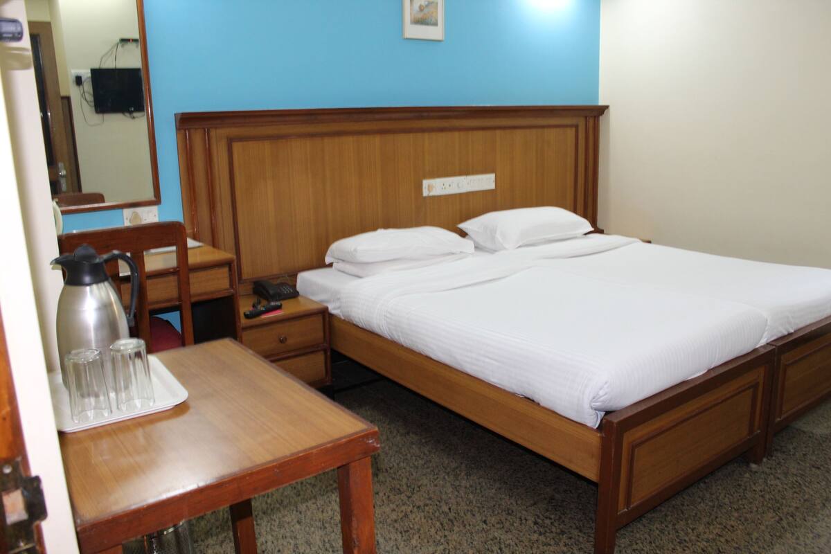 A neatly made bed with white linens, a wooden nightstand with a lamp, a small table with a water pitcher and glasses, and a blue wall with a picture.