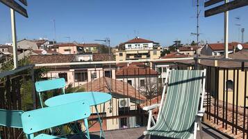 Un balcon avec des chaises turquoise et un fauteuil à rayures, donnant sur un quartier résidentiel avec des maisons et des antennes paraboliques.