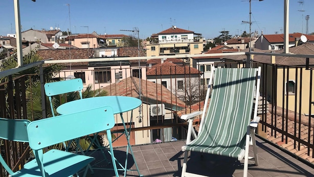 Un balcón con sillas turquesas y una lounge chair a rayas, con vista a una zona residencial con casas y antenas parabólicas.